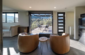 Ein modernes Wohnzimmer mit einer Couch, zwei Sesseln, einem kleinen runden Tisch und einem großen Fenster mit Blick auf eine Terrasse und eine schöne Aussicht.