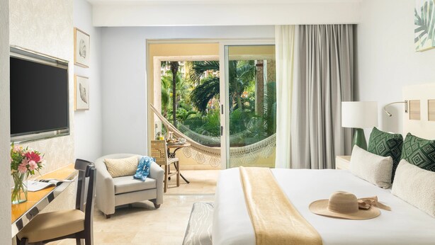 Ein Hotelzimmer mit einem Bett, einem Schreibtisch mit Stuhl, einem Fernseher und Blick auf einen Garten durch eine Glastür.