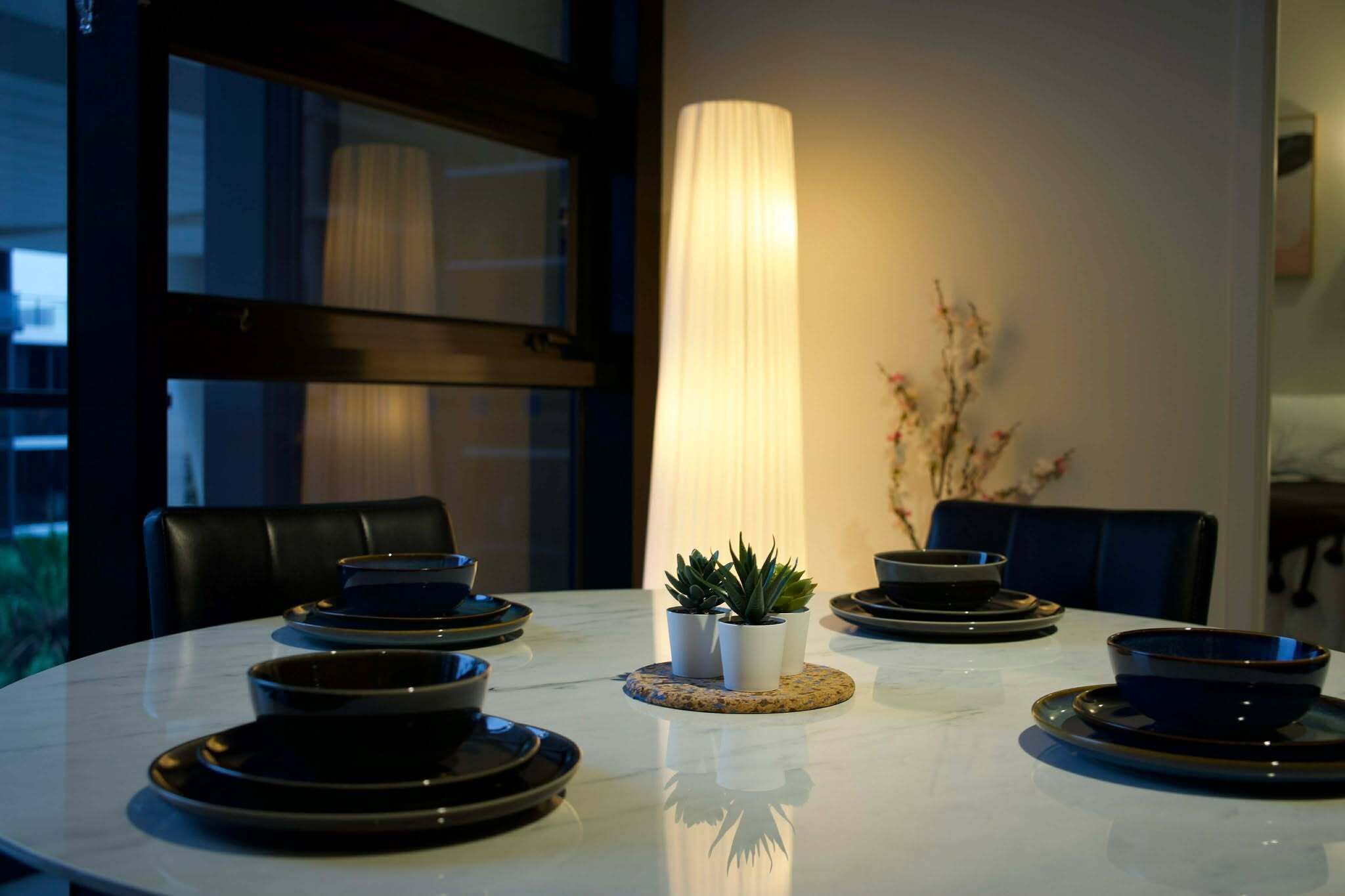 A dining table with black plates and bowls, a tall white lamp, and two potted plants.