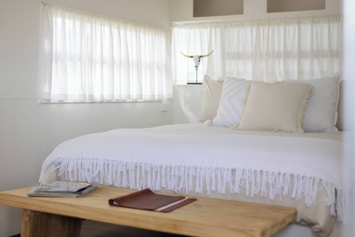 Une chambre à coucher avec un lit, une table de chevet, une lampe et une fenêtre ornée de rideaux légers.
