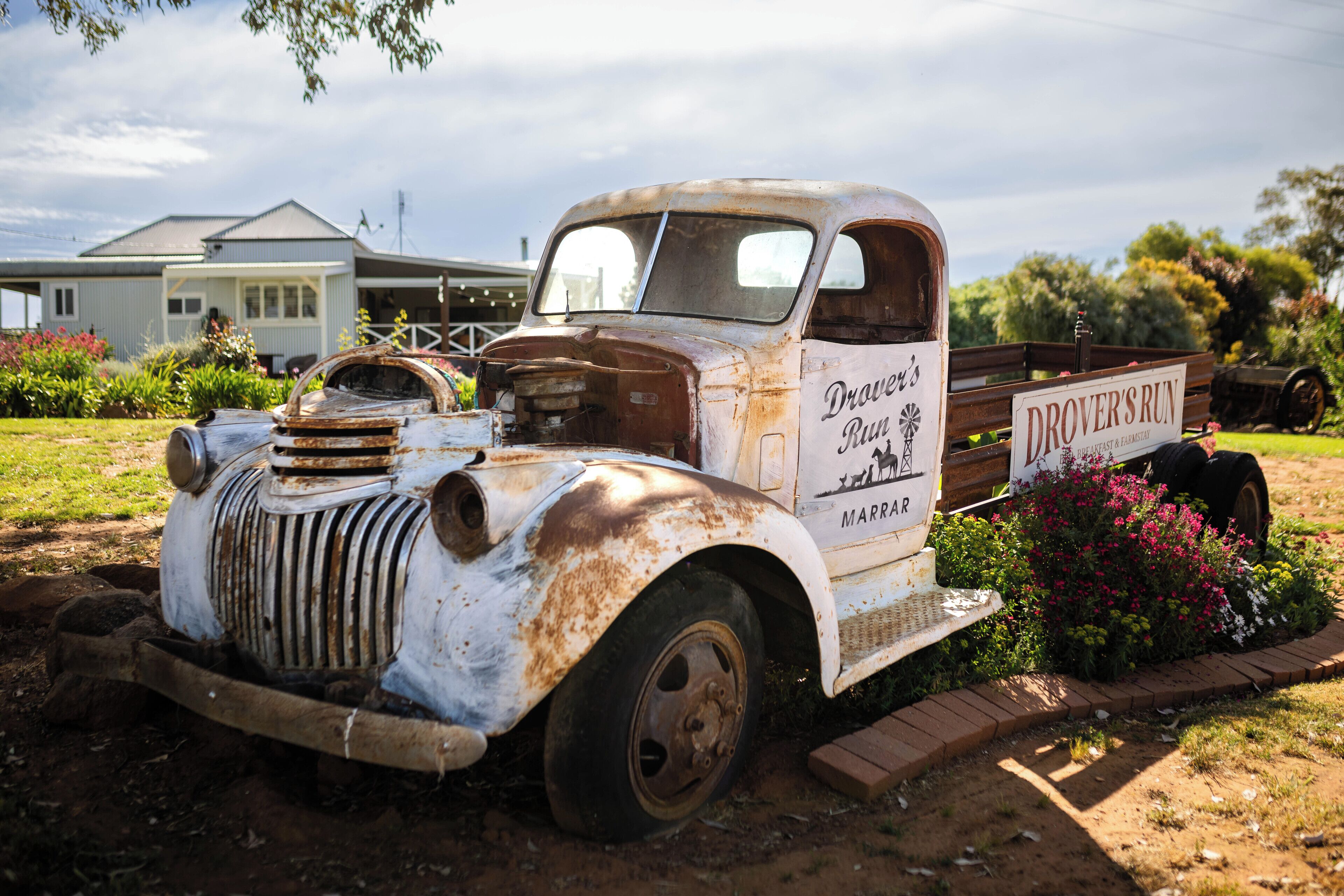 An old, rusted truck with a sign that reads "Drove's Run" parked in a garden with flowers and a house in the background.
