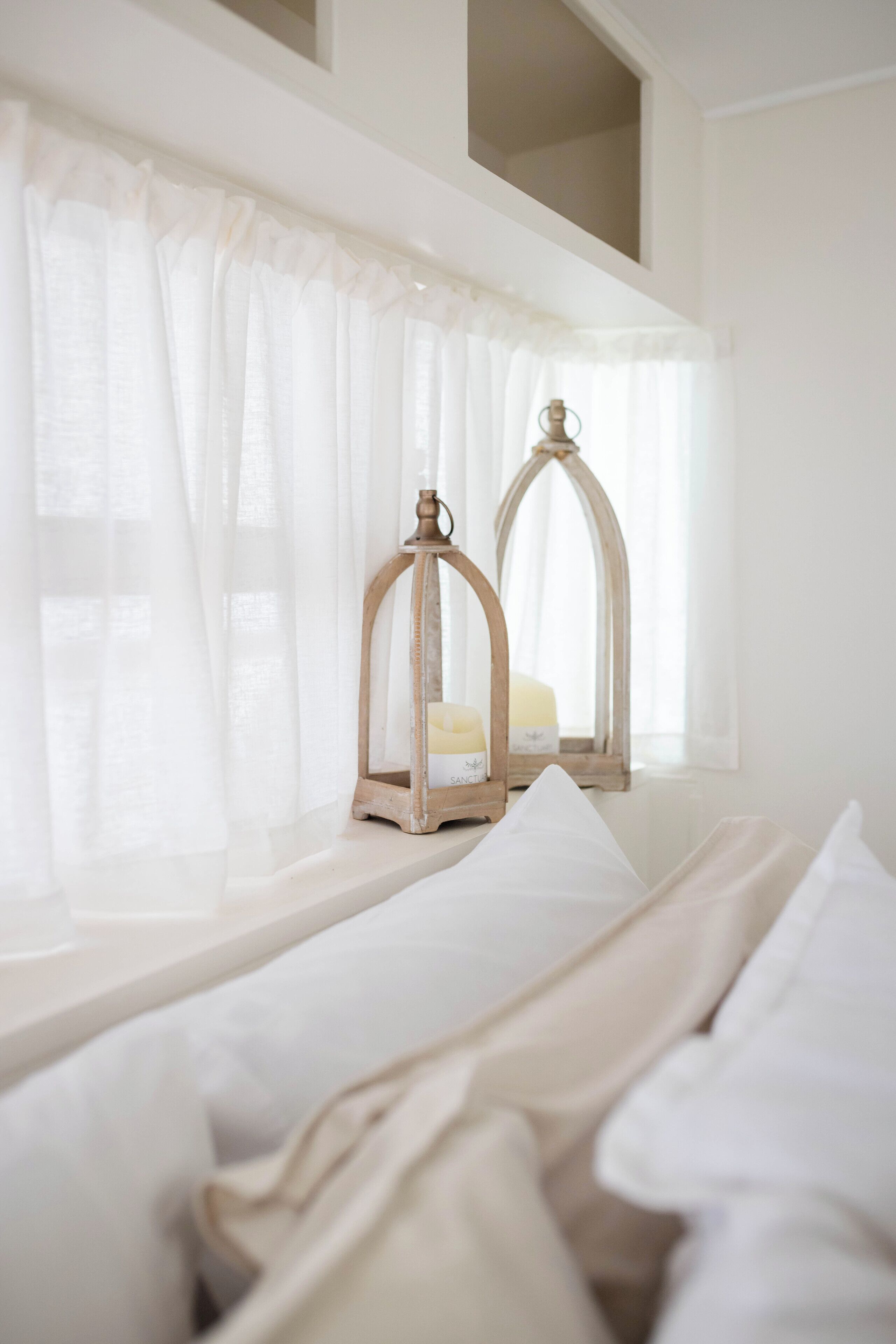 Un dormitorio con cama, cortinas blancas y dos faroles de madera en el alféizar de una ventana.