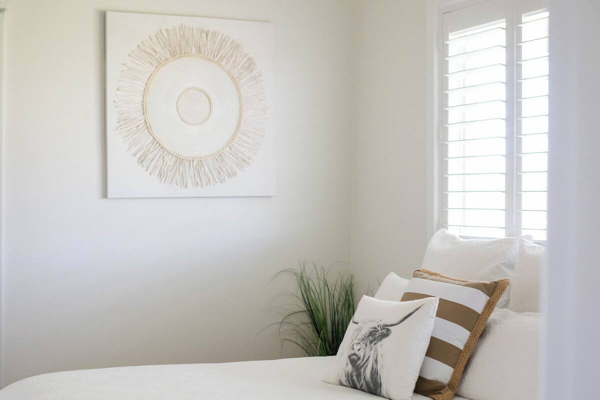 Un lit bien fait, recouvert d’une couvre-lit blanche et agrémenté d’oreillers, une œuvre d’art abstraite accrochée au mur et une fenêtre avec des stores.