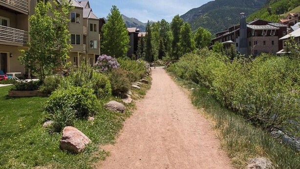 En støvet sti der løber gennem et boligområde med huse på begge sider, omgivet af grønt og med bjerge i baggrunden.