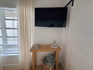 Quarto com televisão na parede, mesa de consola de madeira com um livro e difusor de vime, cesto com manta e janela com persianas.