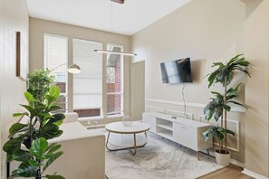 A modern living room with a white sofa, a round coffee table, a flat-screen TV mounted on the wall, and several potted plants. - Modern 4B Retreat with Pool in Frisco (Frisco)