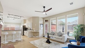 A spacious living area with a sofa, blue chairs, a coffee table, and a dining area with a kitchen in the background. - Modern 4B Retreat with Pool in Frisco (Frisco)