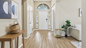 A hallway with a wooden floor, a white door, a framed artwork, a wicker basket, a small table, a potted plant, and a white sofa. - Modern 4B Retreat with Pool in Frisco (Frisco)