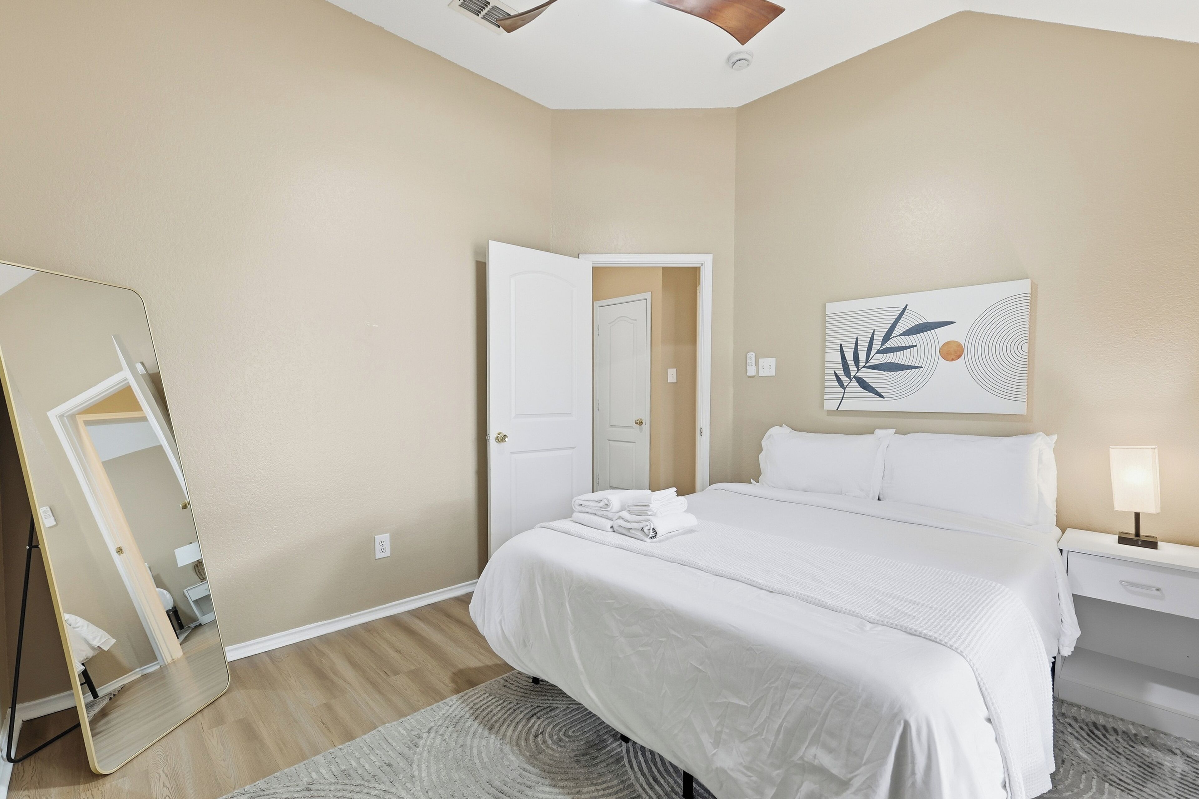 A hotel room with a bed, a nightstand, a lamp, a mirror, and a door.