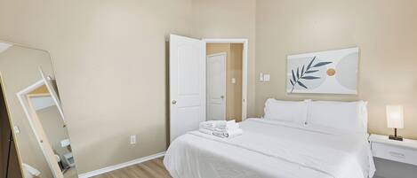 A hotel room with a bed, a nightstand, a lamp, a mirror, and a door.