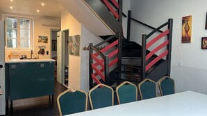 Una cocina moderna con armarios color turquesa, una encimera blanca y un comedor con mesa blanca y sillas turquesa. Una escalera con barandales rojos conduce a un nivel superior.