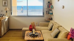 Una sala de estar con un ventanal grande que da a una playa, un sofá, una mesita de centro y una estantería.
