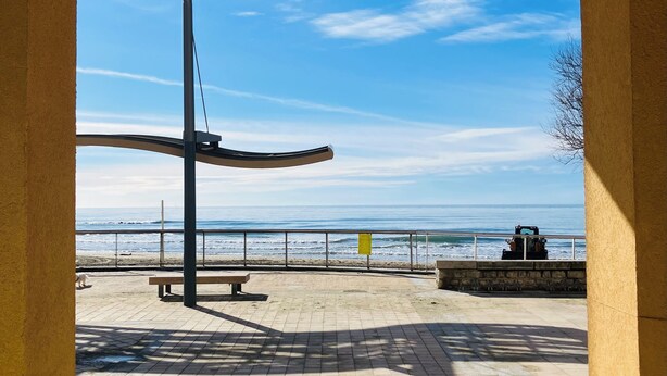 Un sendero pavimentado con un banco y una escultura de metal, con vistas a una playa que tiene barandilla y una lejana perspectiva del océano.