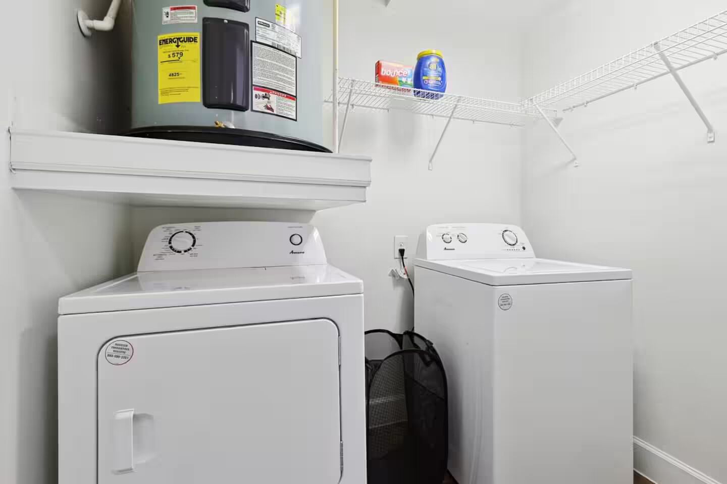 A laundry room with two front-loading washers and dryers, a shelf with cleaning supplies, and a water heater above the dryers.