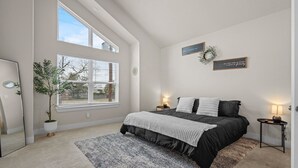 A bedroom with a large window, a bed with a black and white bedspread, a small side table with a lamp, and a potted plant. - Modern 3BR Near RiverWalk•Sleeps 8•Free Parking (San Antonio)