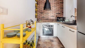 A compact kitchen with white cabinets, a brick backsplash, a yellow shelf with dishes, and a black hanging light fixture. - Bright & Stylish 2 bed Apartment in Trastevere (Roma)