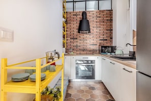 A compact kitchen with white cabinets, a brick backsplash, a yellow shelf with dishes, and a black hanging light fixture. - Bright & Stylish 2 bed Apartment in Trastevere (Roma)