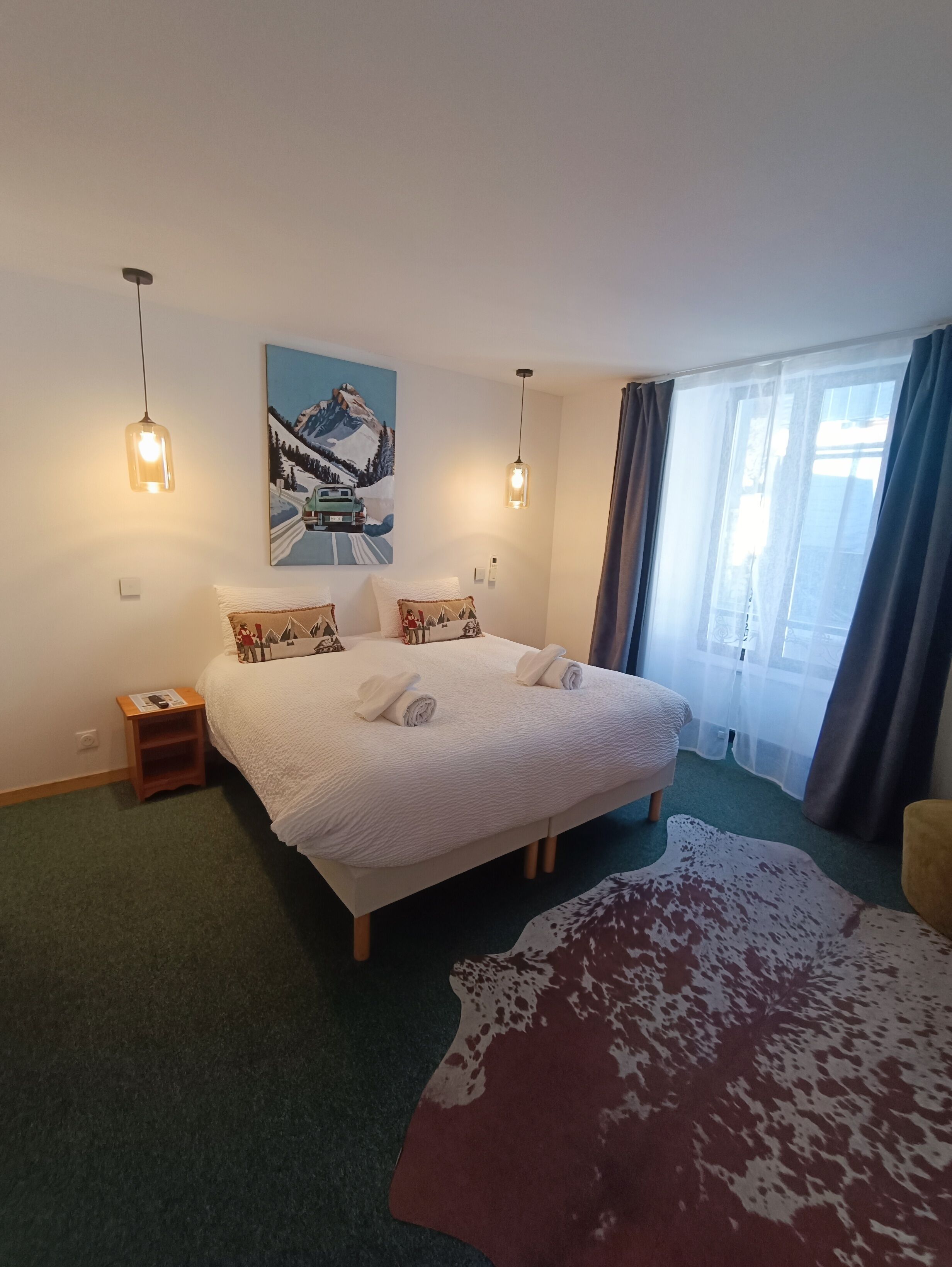 Habitación de hotel con una cama grande, dos lámparas de noche, un cuadro en la pared, una mesita de madera pequeña y un ventanal con cortinas.