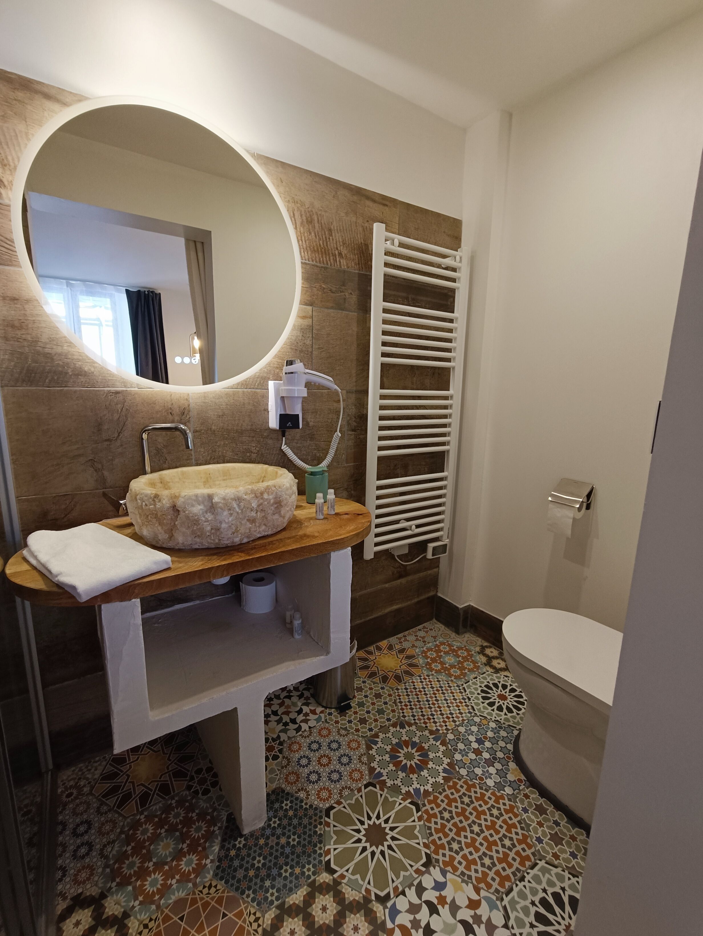 Une salle de bain avec un miroir rond, un meuble-lavabo en bois, une serviette, des toilettes et un carrelage à motifs.