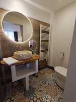 A bathroom with a round mirror, a wooden vanity, a towel, a toilet, and a patterned floor.