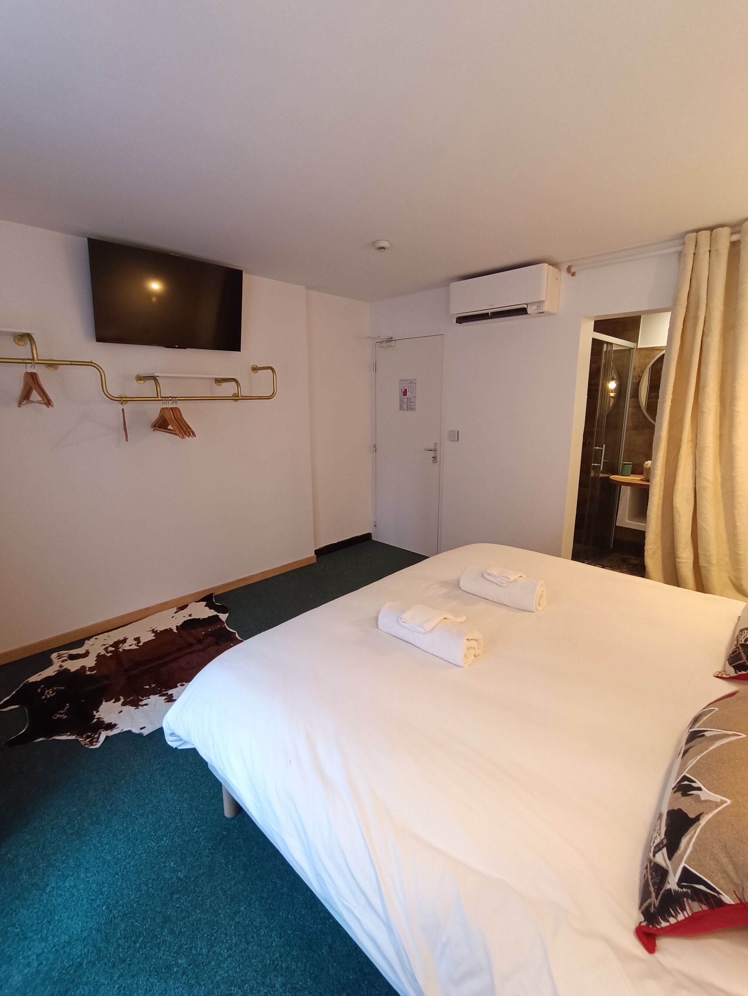 Habitación de hotel con cama, televisor montado, perchero y puerta que da a un baño.