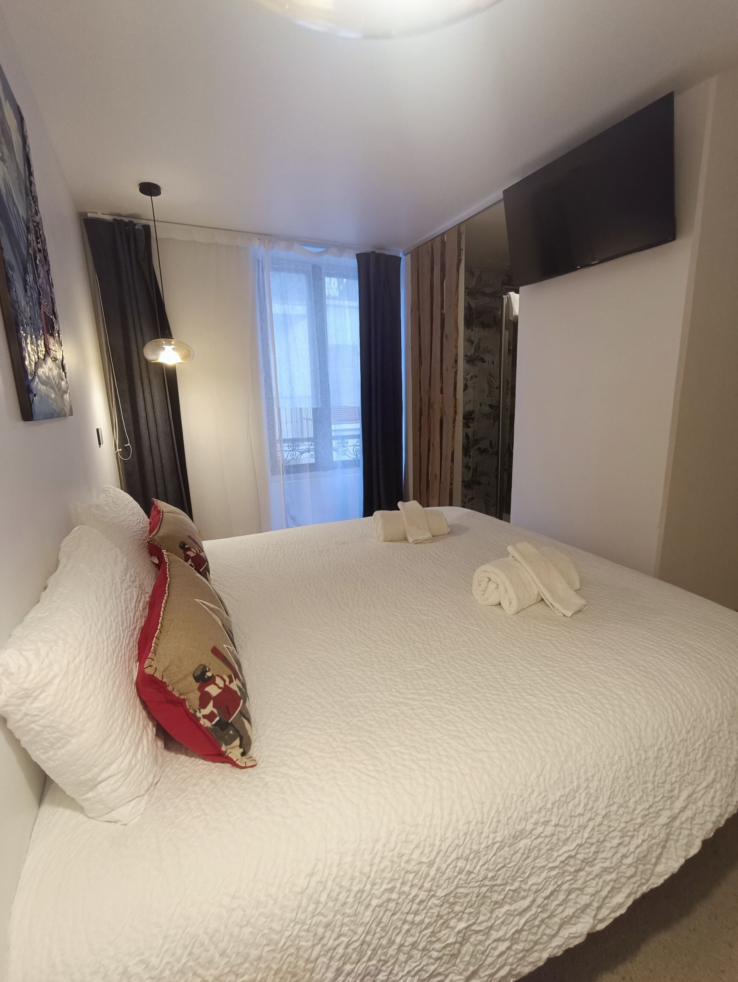 A hotel room with a large bed, a flat-screen TV mounted on the wall, a window with curtains, and a painting on the wall.