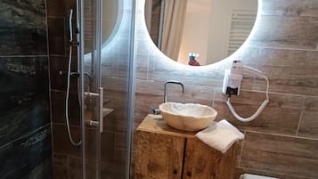 A bathroom with a glass shower enclosure, a wooden vanity with a round mirror, a sink, and a toilet.
