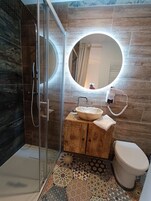 A bathroom with a glass shower enclosure, a wooden vanity with a round mirror, a sink, and a toilet.