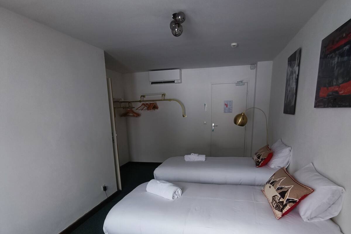 A hotel room with two beds, a bedside table, a lamp, a coat rack, and a door.
