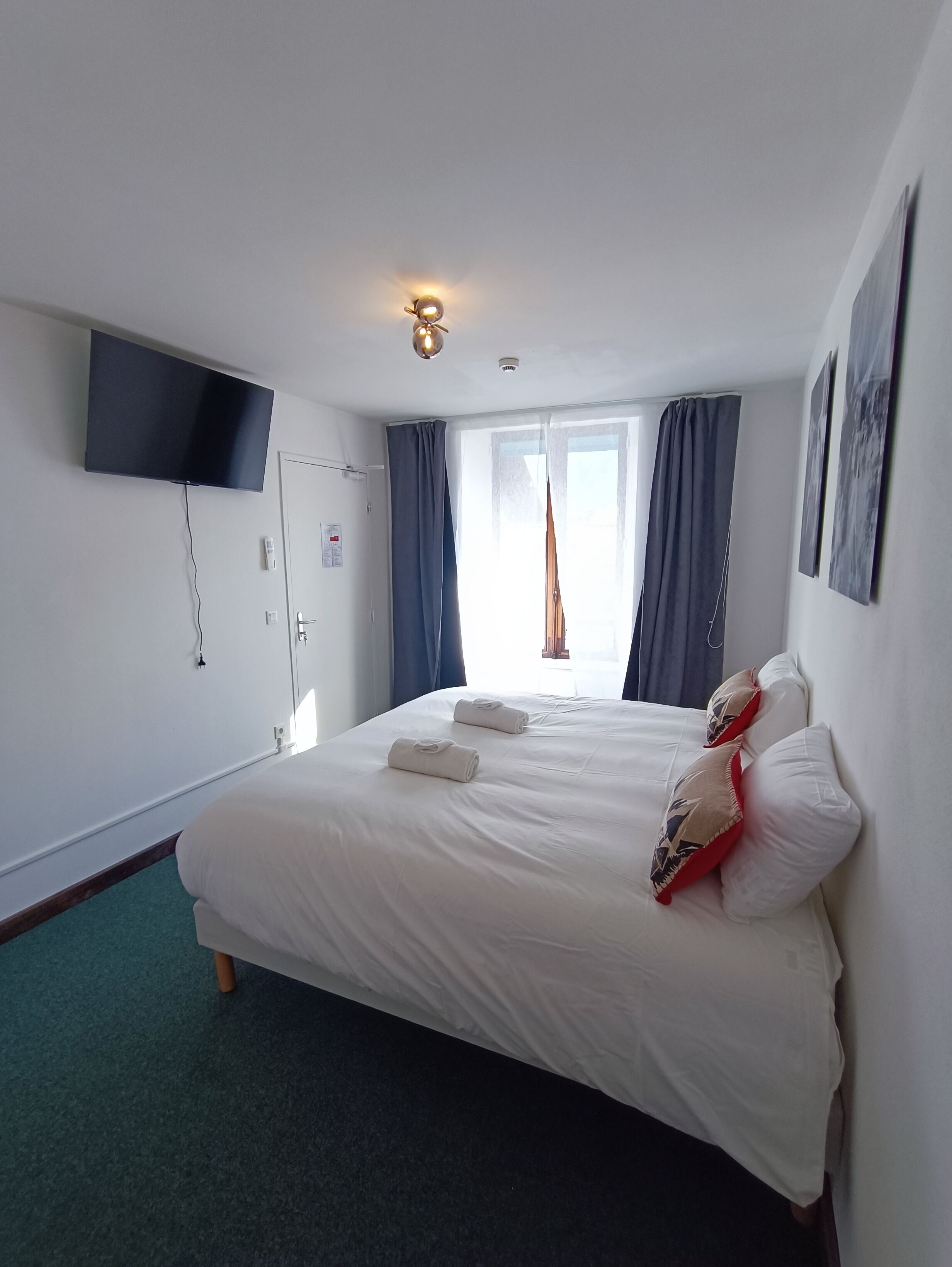 A hotel room with a bed, a flat-screen TV mounted on the wall, a window with curtains, and a ceiling light.