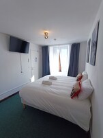 Ein Hotelzimmer mit Bett, einem flachbildfernseher an der Wand, einem Fenster mit Vorhängen und einer Deckenleuchte.