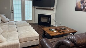 A living room with a sectional sofa, a fireplace, a coffee table, and a flat-screen TV mounted above the fireplace. - Simple Comfort 2-bedroom home 4.5 miles from the Augusta National Golf Course! (Augusta)