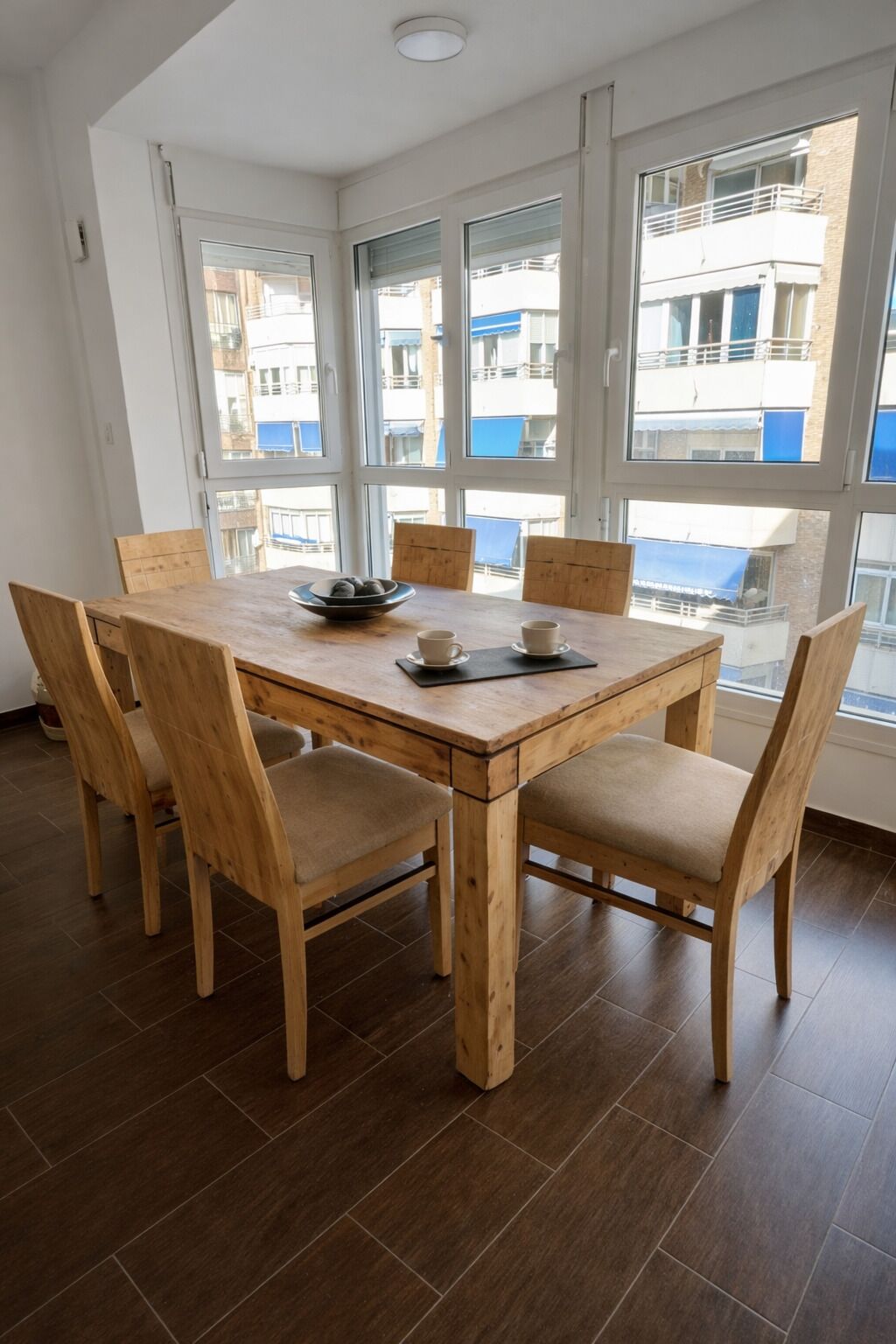 A dining area with a wooden table and chairs, large windows with a view of buildings, and a tiled floor.