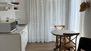 A compact kitchen with white appliances, a dining area with a table and chairs, and a window with sheer curtains. - Self-Contained Studio Apartment – Macedon Ranges, Riddells Creek (Riddells Creek)