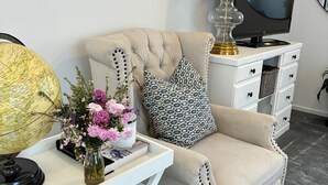 A well-lit room with a beige armchair, a small desk with a globe and flowers, a lamp, and a TV on a white dresser. - Self-Contained Studio Apartment – Macedon Ranges, Riddells Creek (Riddells Creek)