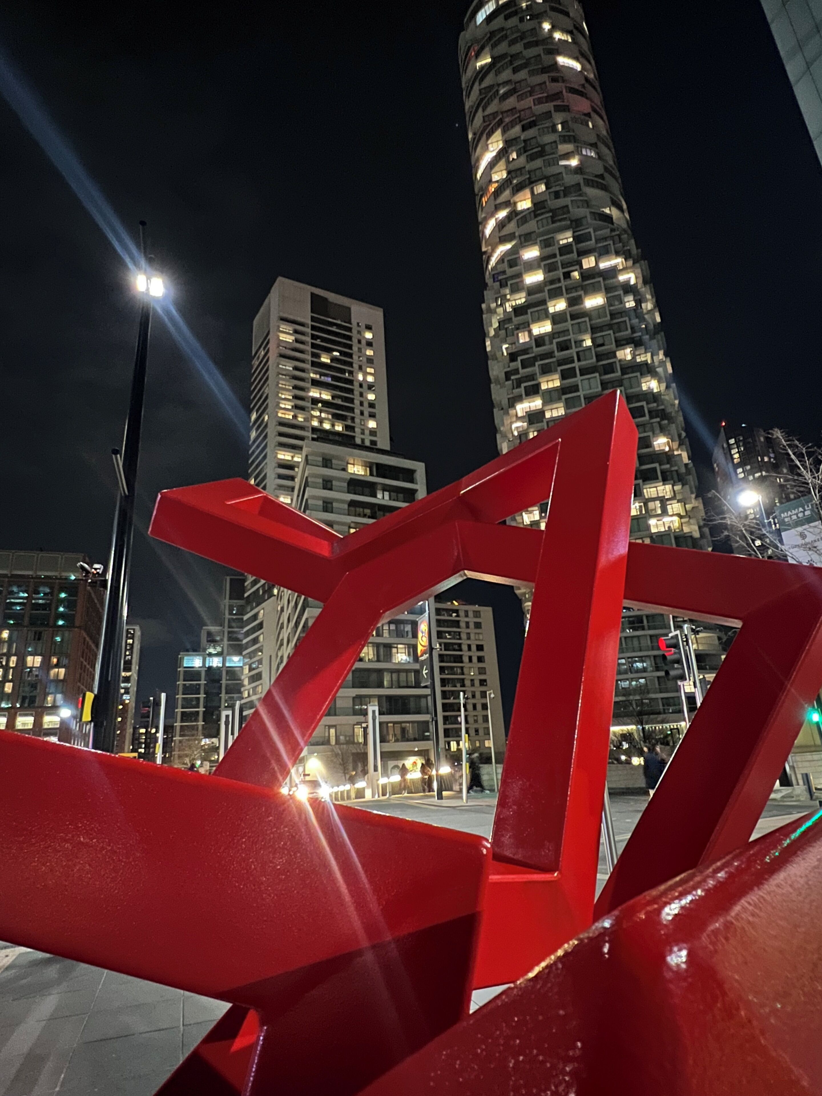 A nighttime cityscape with illuminated buildings and a prominent red sculpture in the foreground.