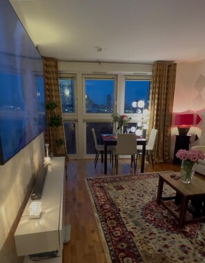 Ein modernes Wohnzimmer mit Flachbildfernseher, einer Essecke mit Stühlen und Tisch sowie großen Fenstern mit Blick auf die Stadt.