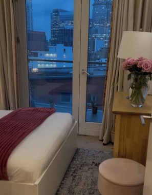 Ein Hotelzimmer mit Bett, einem kleinen Tisch mit Blumenvase, einem Sessel und Blick auf eine Stadt durch eine Glasdoor.
