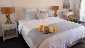 Ein ordentlich bezogenes Bett mit grau-weißer Bettdecke, zwei Nachttische mit Lampen, ein Holzstuhl und ein Schreibtisch mit Stuhl.