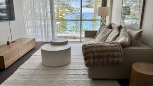 Ein modernes Wohnzimmer mit großem Fenster und Meerblick, einem runden weißen Hocker, einem beige farbenen Sofa mit Decke und einer Holzkommode.