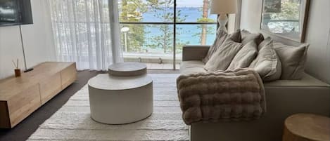 A modern living room with a large window offering a view of the sea, a round white ottoman, a beige sofa with a throw blanket, and a wooden sideboard.