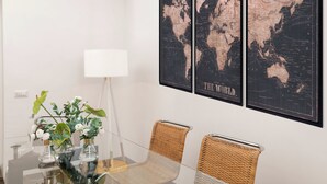 Un coin repas avec une table en verre, des chaises en osier et un vase rempli de fleurs. Deux cartes du monde encadrées sont accrochées au mur.
