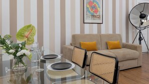 Un salon moderne avec une table à manger en verre, un canapé beige agrémenté de coussins jaunes, une œuvre d’art abstraite encadrée et une lampe sur pied.