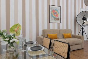 Ein modernes Wohnzimmer mit einem Glastisch, einem beigefarbenen Sofa mit gelben Kissen, einem gerahmten abstrakten Kunstwerk und einer Stehlampe.