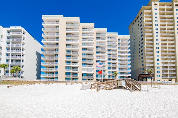 A beachfront hotel with multiple balconies, a sandy beach in front, and a wooden walkway leading to the entrance. - Oceanfront 3BR Corner Unit | Wraparound Balcony | Sleeps 8 (Orange Beach)