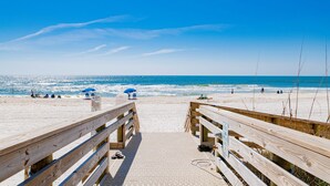 Une promenade en bois mène à une plage de sable fin, sous un ciel bleu et dégagé.