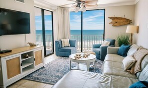 A living room with a TV, sofas, a coffee table, and a view of the ocean. - Oceanfront 3BR Corner Unit | Wraparound Balcony | Sleeps 8 (Orange Beach)
