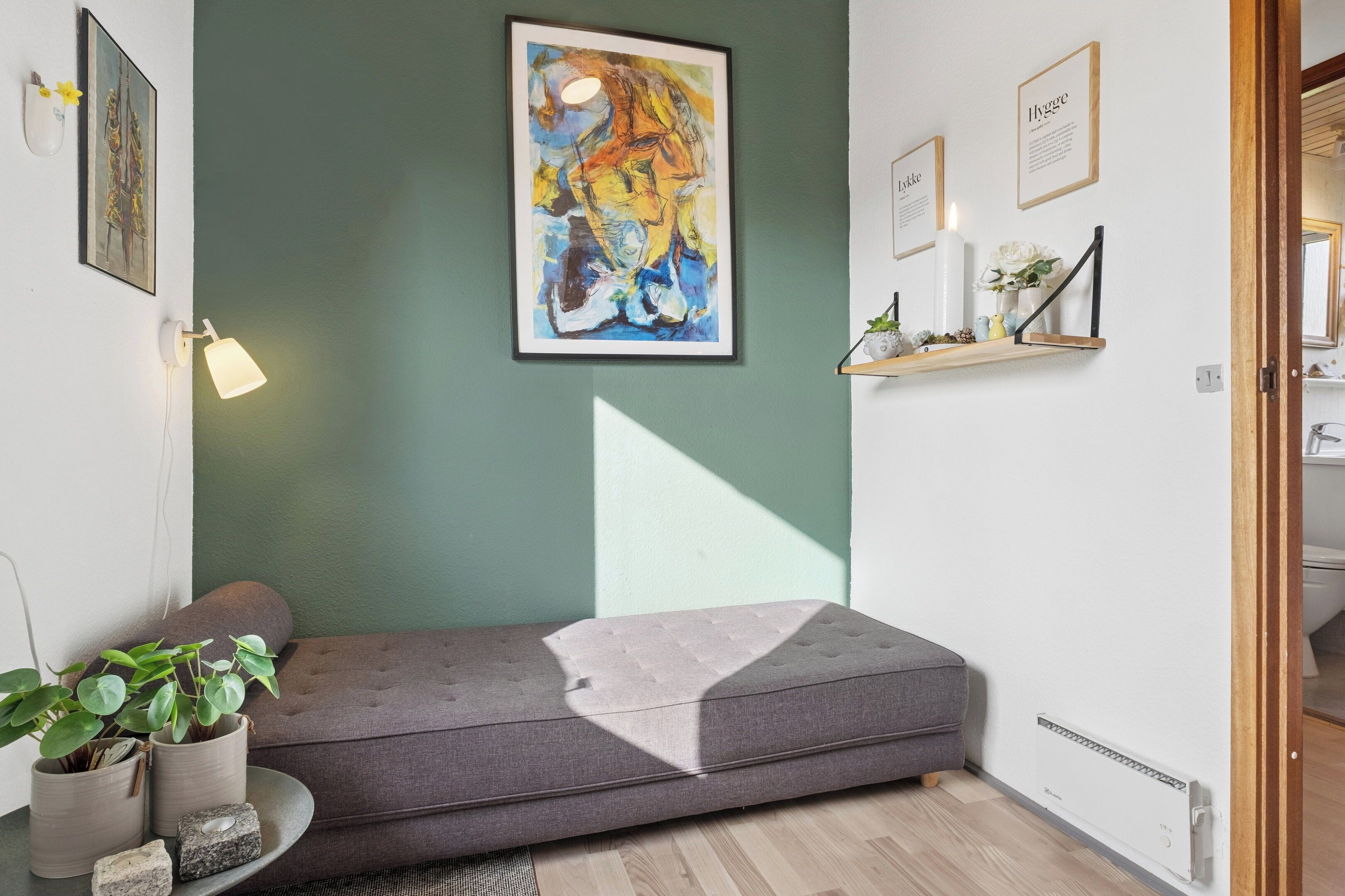 Una habitación con un sofá gris, una pared verde, un cuadro abstracto enmarcado, un estante con adornos y un radiador.