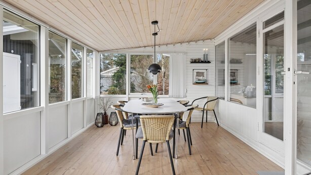 Una habitación amplia con una mesa redonda, sillas de mimbre, una lámpara colgante y ventanas grandes.