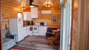 A compact kitchen with white cabinets, a microwave, a stove, and a refrigerator. There is a gray sofa and a wooden dining table with chairs. - Gorgeous 2-bedroom cottage in charming Hammondsport awaits you (Hammondsport)